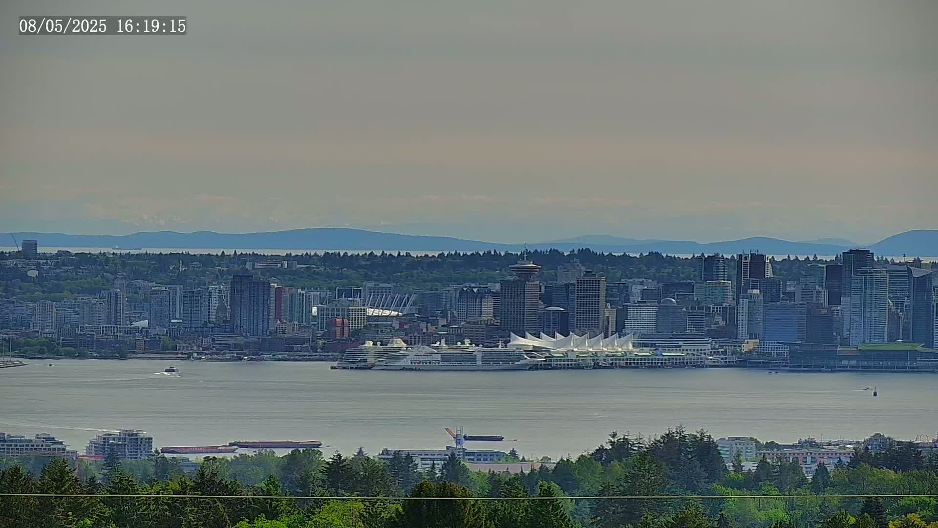 A hazy, distant view of a city skyline with a cruise ship docked near a large white structure, all situated on a body of water with mountains visible in the background.
