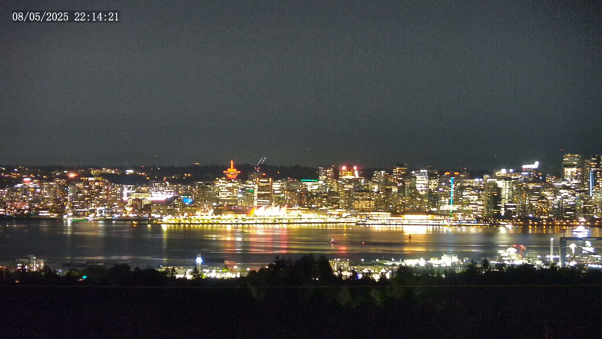 A brightly lit city skyline across a dark body of water is seen from a distance at night.
