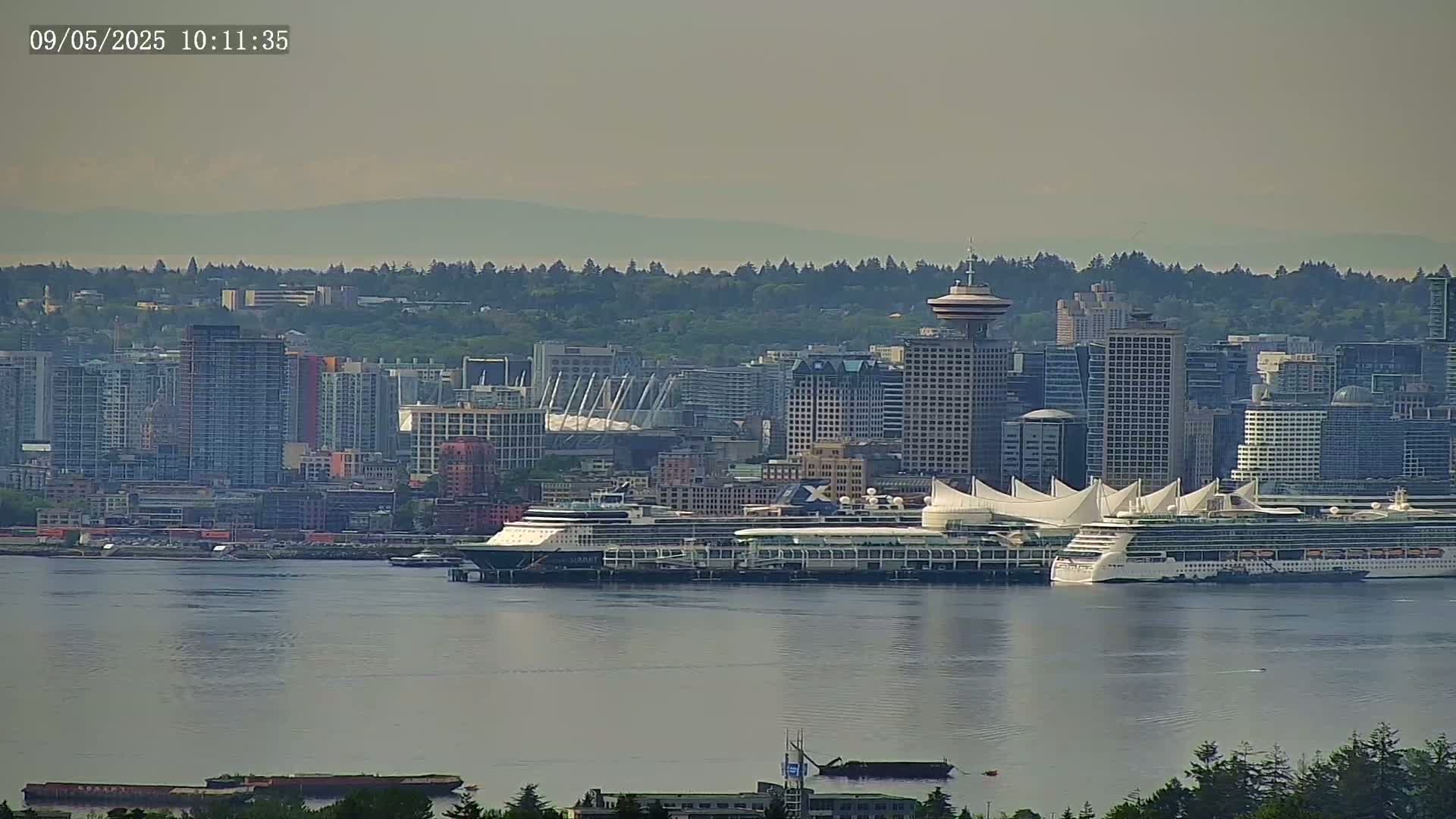 Two cruise ships are docked at a city harbor on a hazy day, with a cityscape of high-rises and a stadium visible in the background.