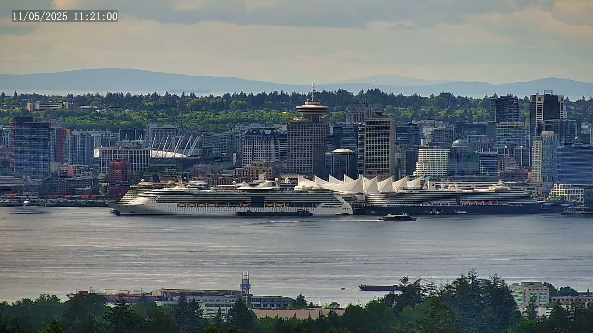 A cruise ship is docked in a city harbor next to a large white-roofed building, with a cityscape and distant mountains visible under a mostly cloudy sky.