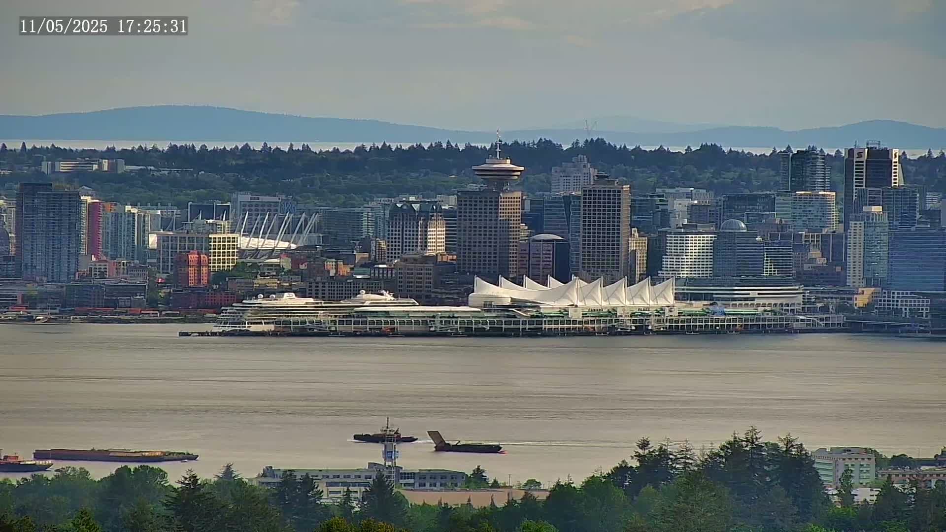 A distant view of a city skyline with a cruise ship docked near a large white-tented structure, under partly cloudy skies.