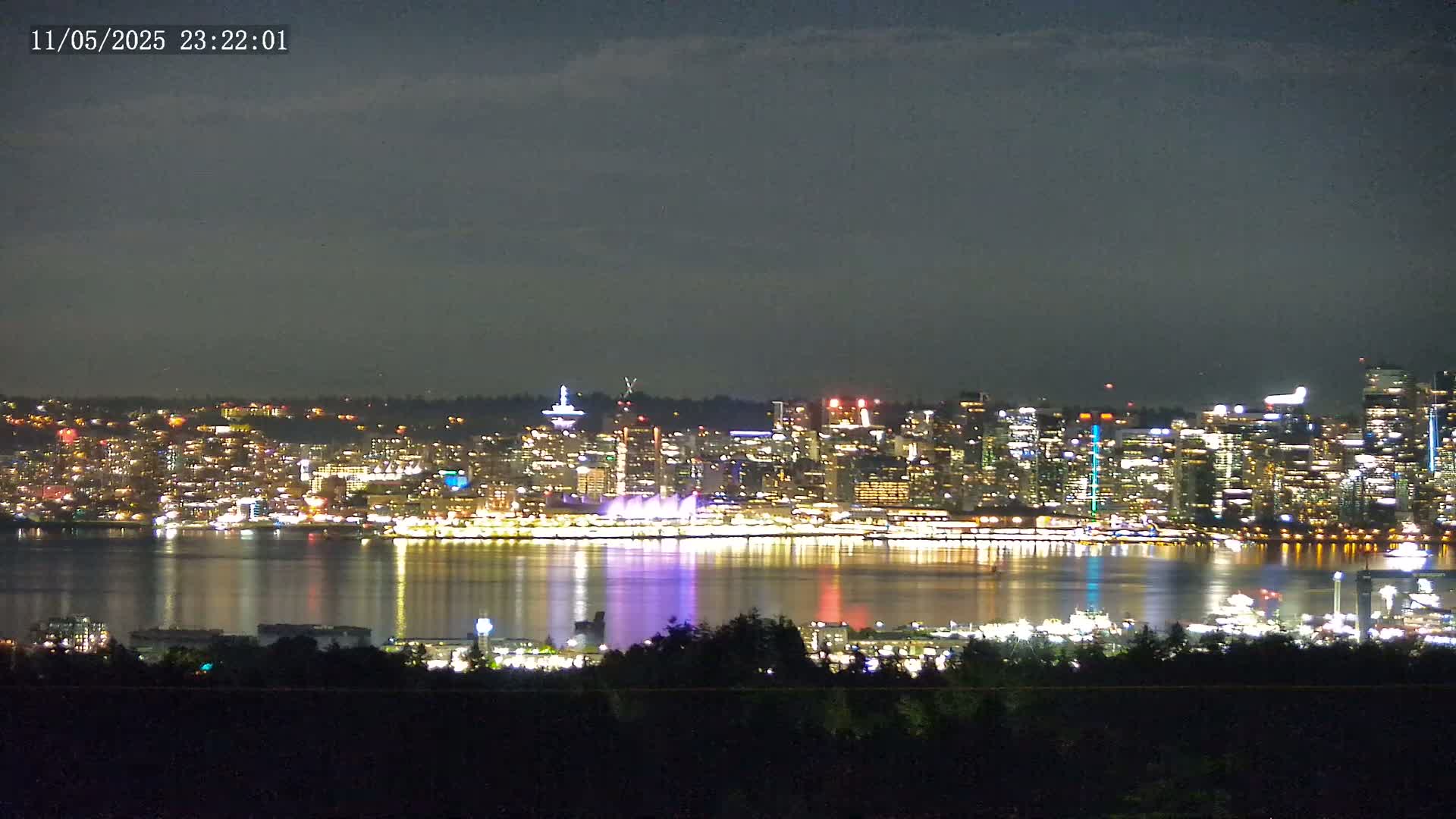 A brightly lit city skyline is seen across a calm body of water at night under a mostly clear sky.