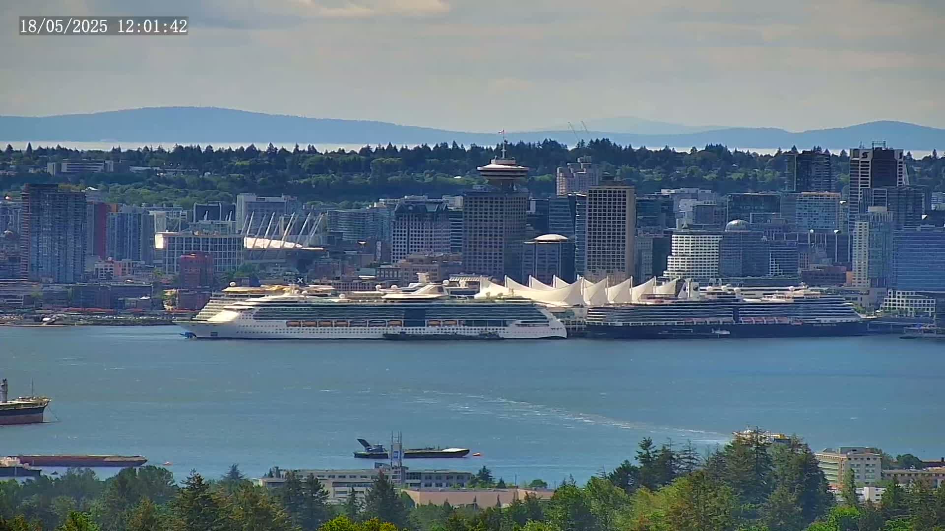 A large cruise ship is docked in a city harbor under partly cloudy skies, with a cityscape and mountains visible in the background.