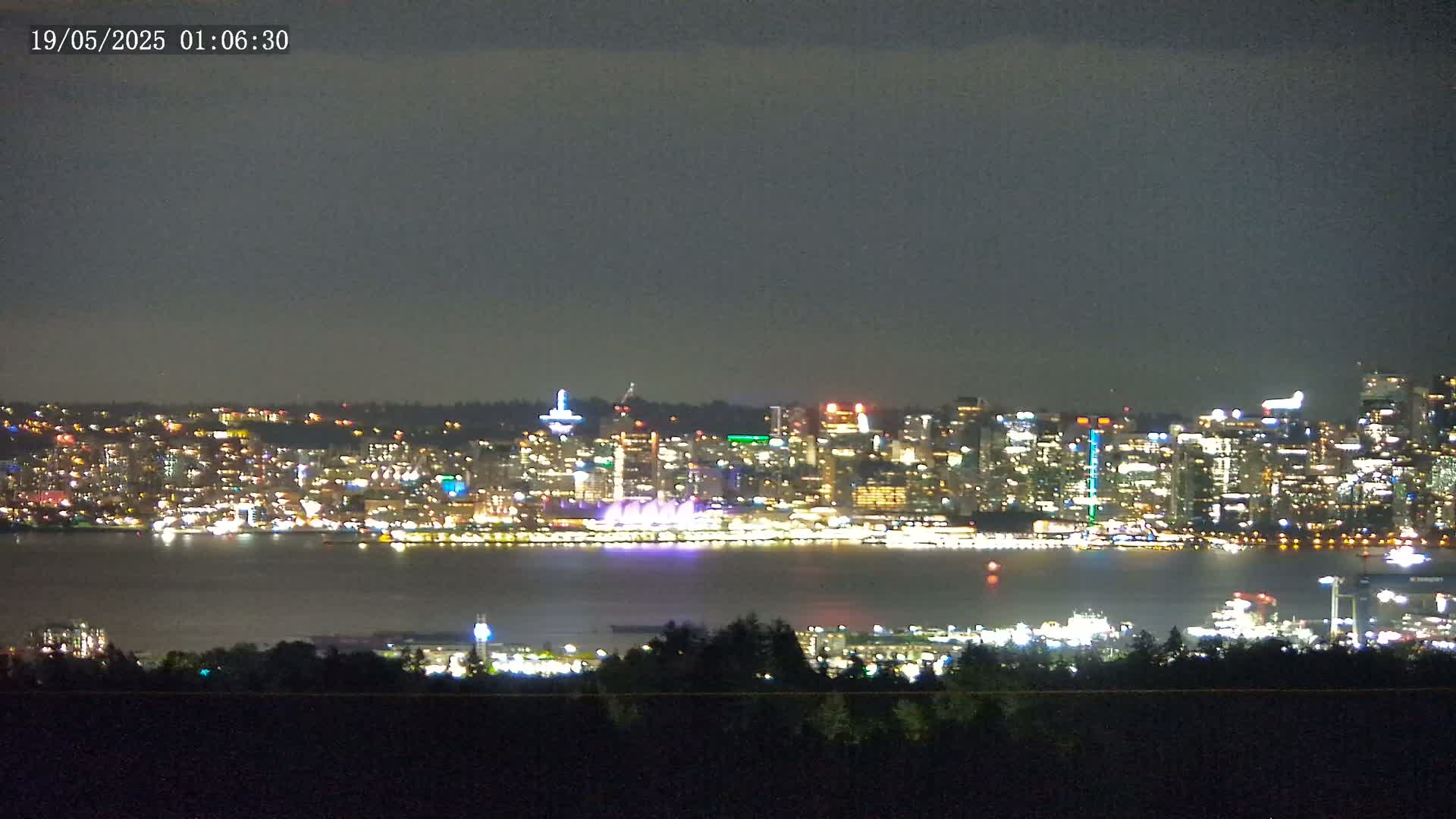 A brightly lit city skyline is seen across a dark body of water at night, under a mostly cloudy sky.