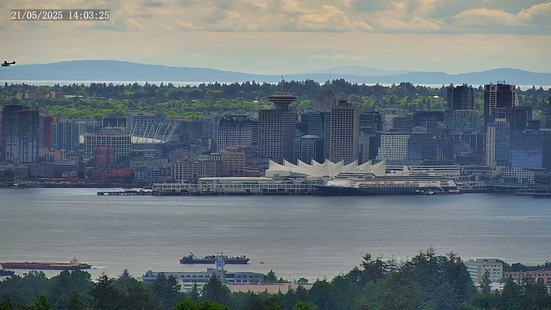 A partly cloudy day shows a cityscape with a large cruise ship docked near a convention center, all situated on a body of water with a distant mountain range visible in the background.