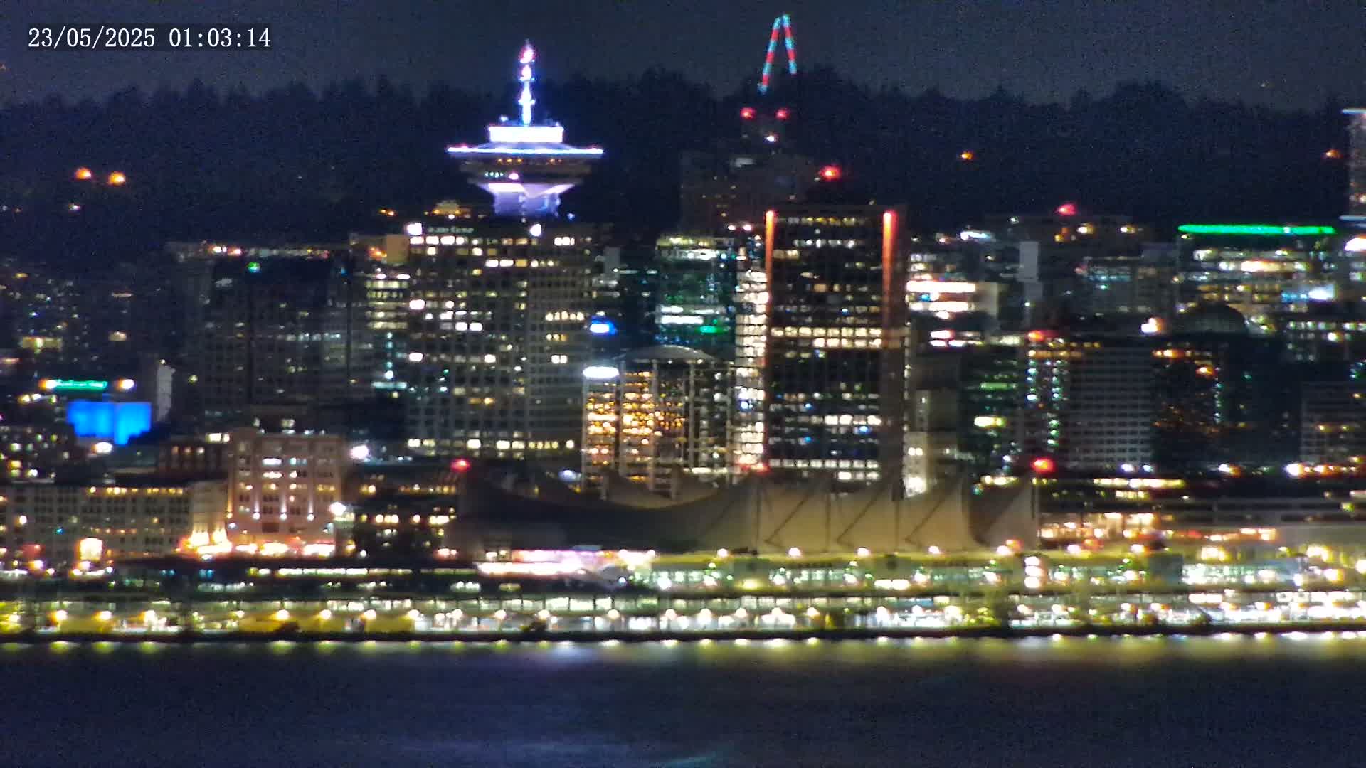 A brightly lit city skyline at night is seen across a dark body of water under a clear, dark sky.