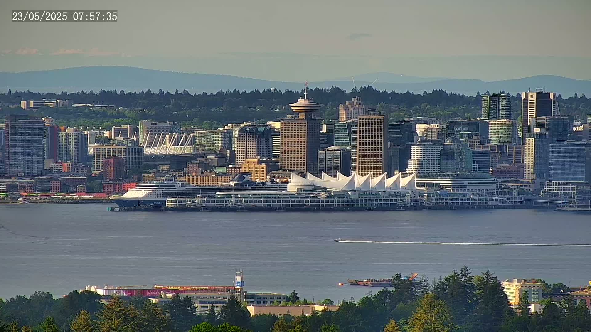 A distant view of a city skyline on a calm, hazy day, featuring a cruise ship docked near a large convention center, with mountains visible in the background.