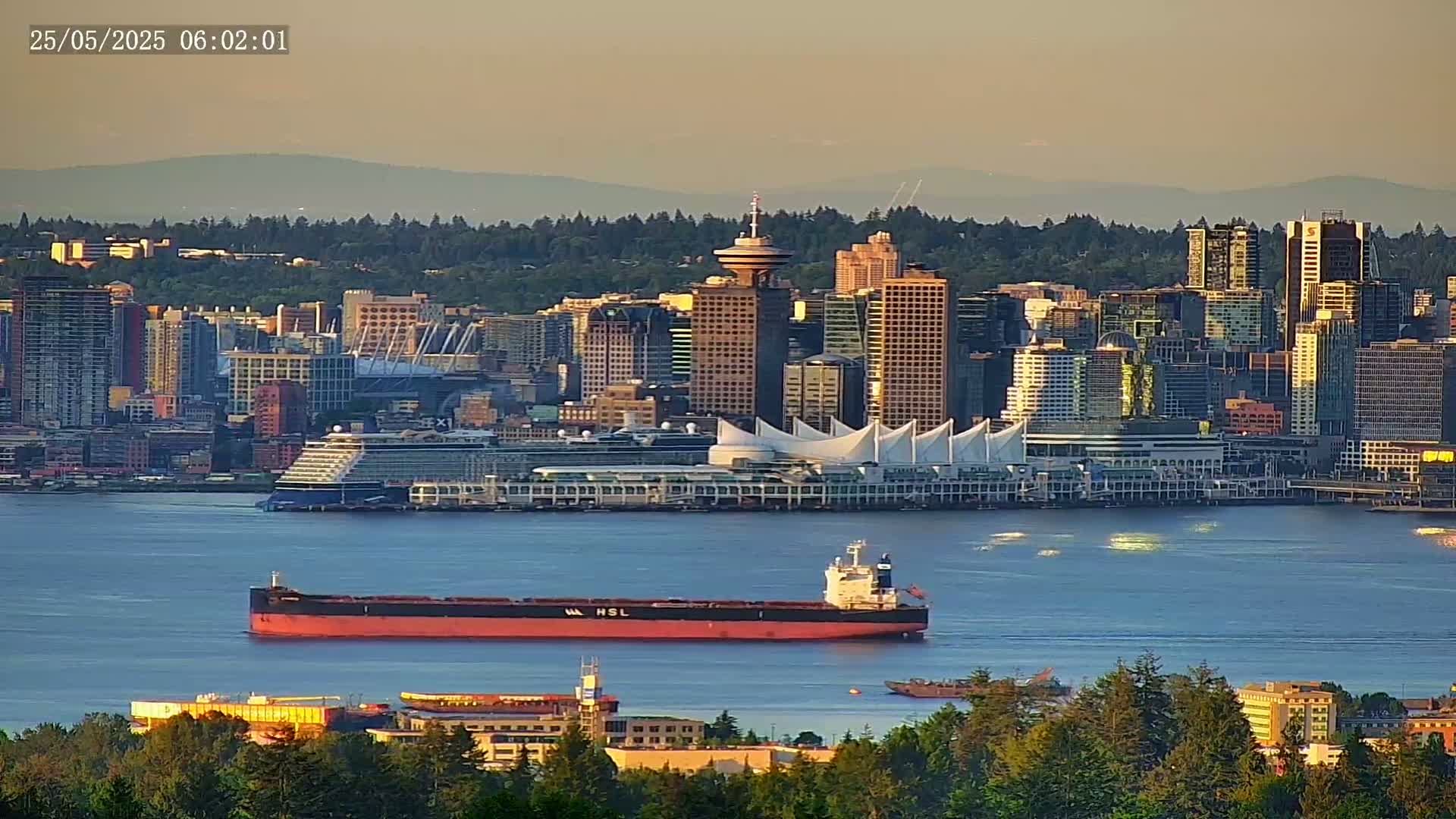 A large cargo ship is in the water near a city skyline at sunrise, with calm water and mountains in the background.