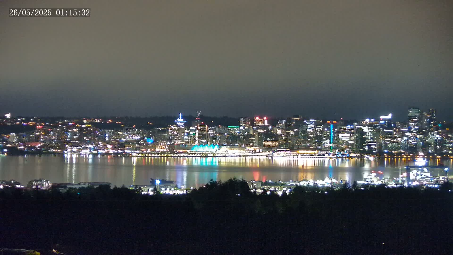 A brightly lit city skyline is reflected in a calm body of water under a dark, overcast night sky.