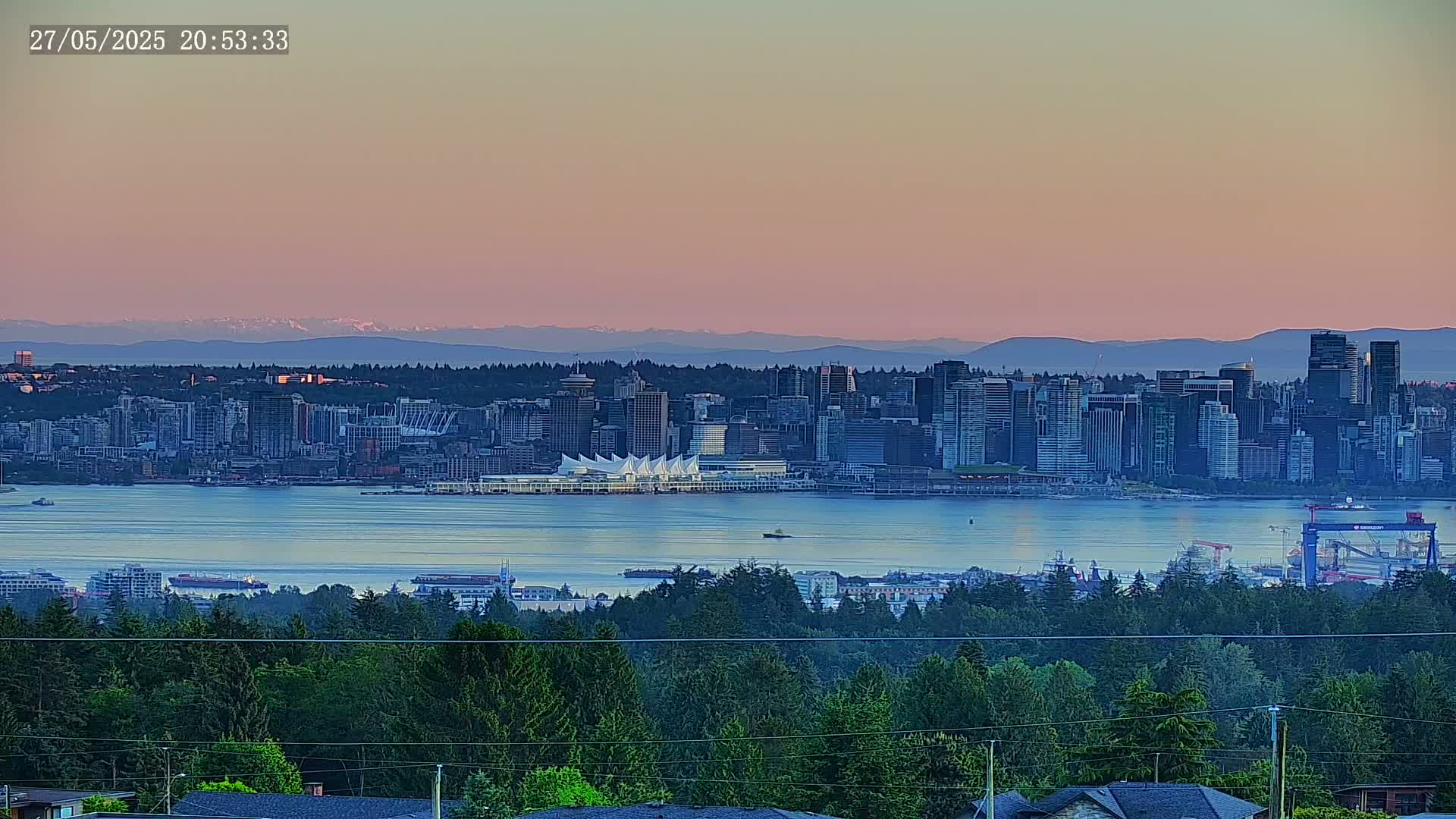 A city skyline, situated on a body of water, is viewed from a distance under a pink and purple sunset sky, with mountains visible in the far background.