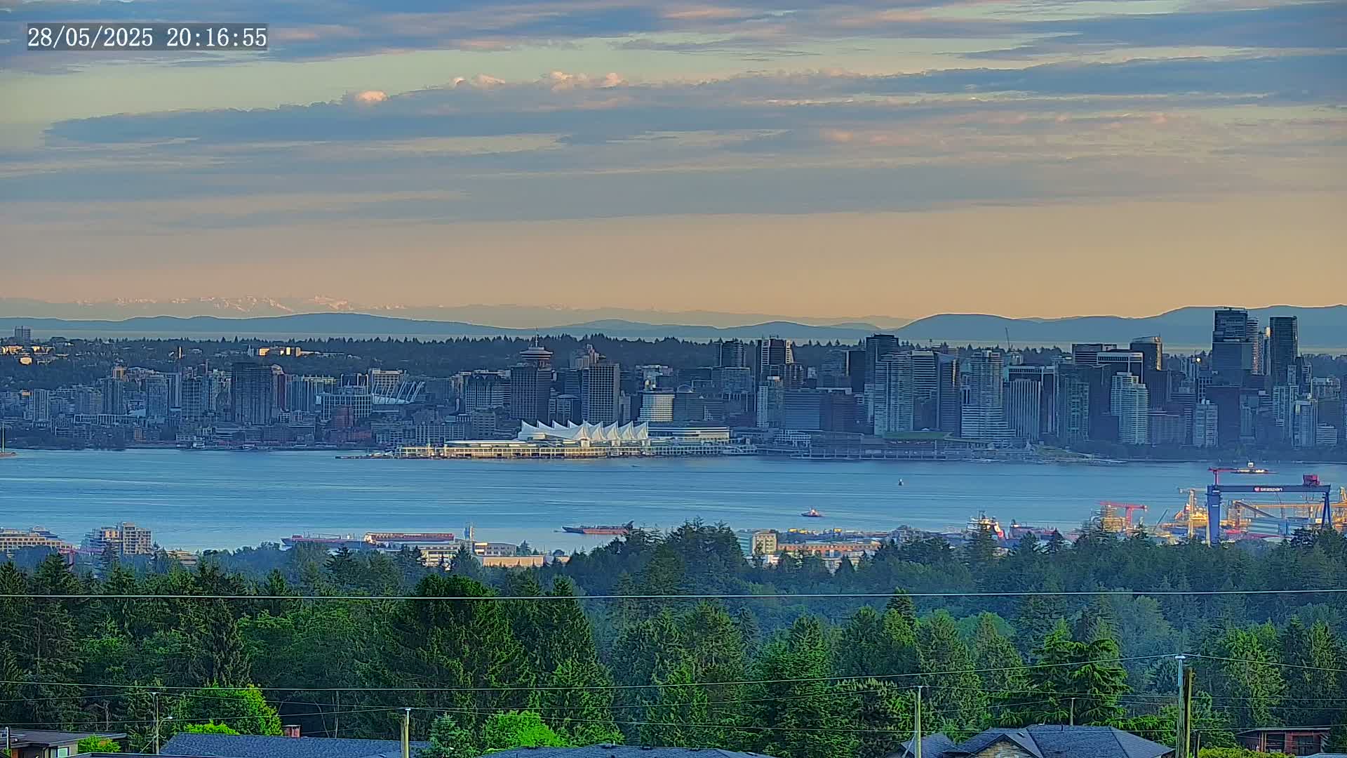 A city skyline sits on a body of water under a partly cloudy sunset sky, viewed from a distance across a forest.