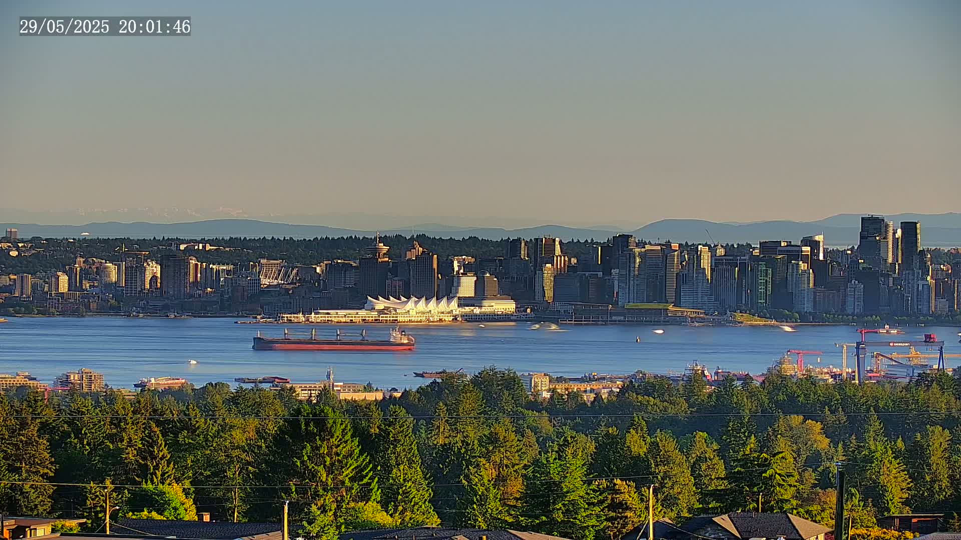 A distant cityscape with skyscrapers and a large white building sits across a calm body of water from a vantage point covered in lush green trees under a clear sky, mountains visible in the far background.