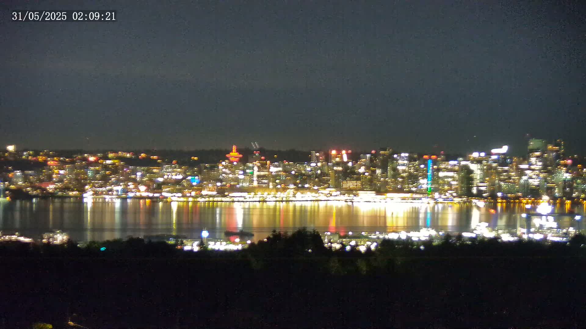 A brightly lit city skyline at night is reflected in calm water, viewed from across a body of water under a dark sky.