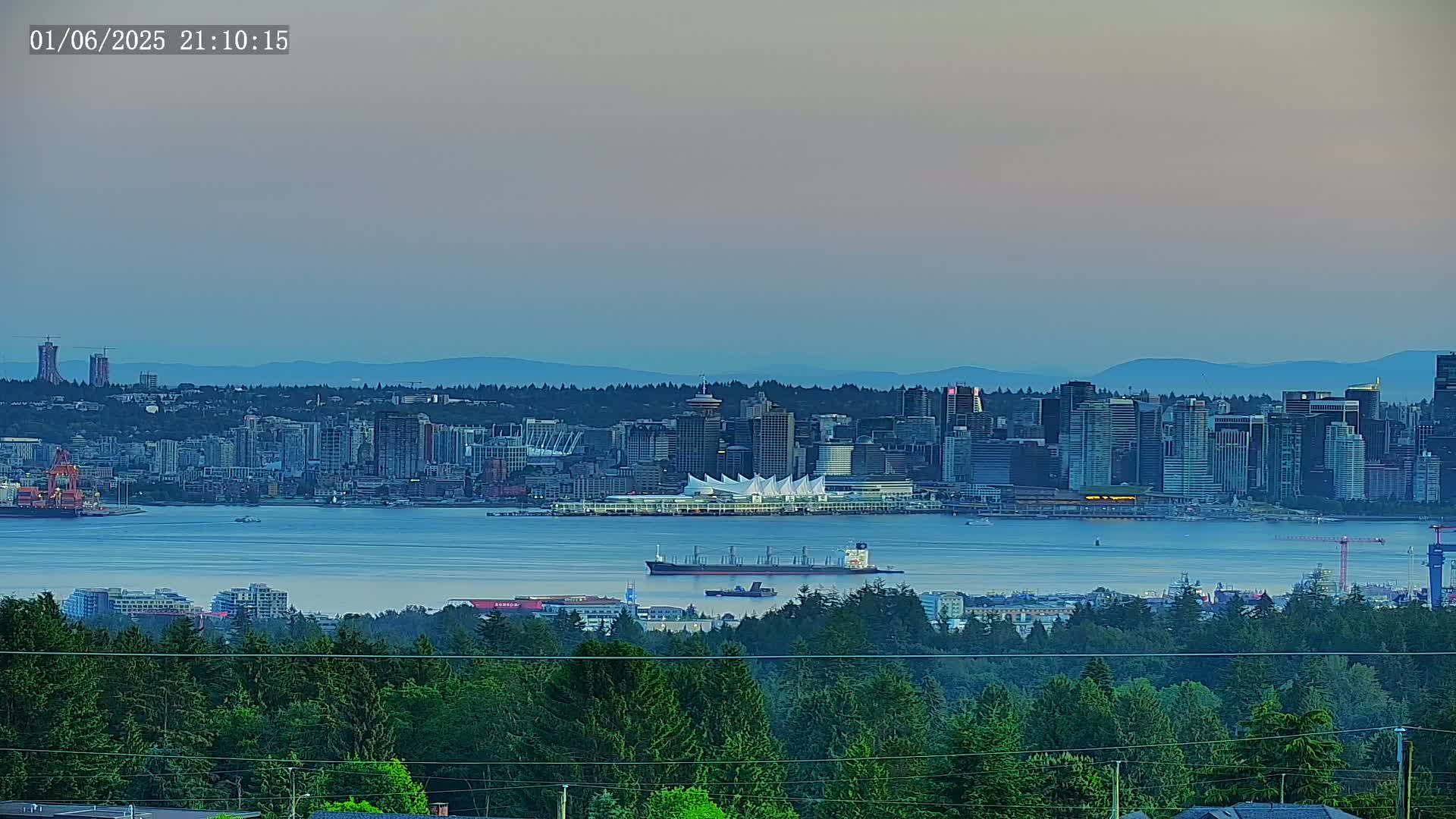 A large city skyline is seen across a calm body of water from a vantage point beyond a forest, under a dusky, twilight sky.
