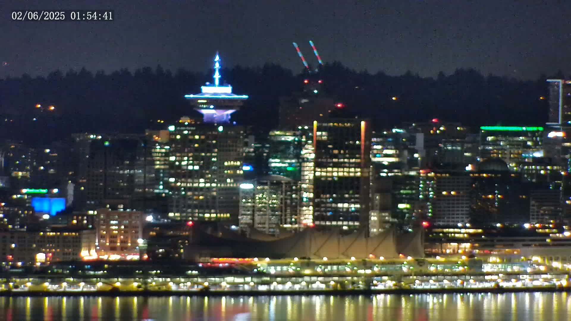 A brightly lit city skyline at night is reflected in calm water under a dark sky.