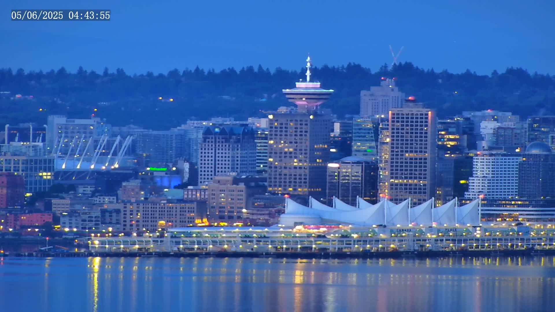 A nighttime cityscape with illuminated buildings, a harbor, and a prominent tower, reflected in calm water under a twilight sky.