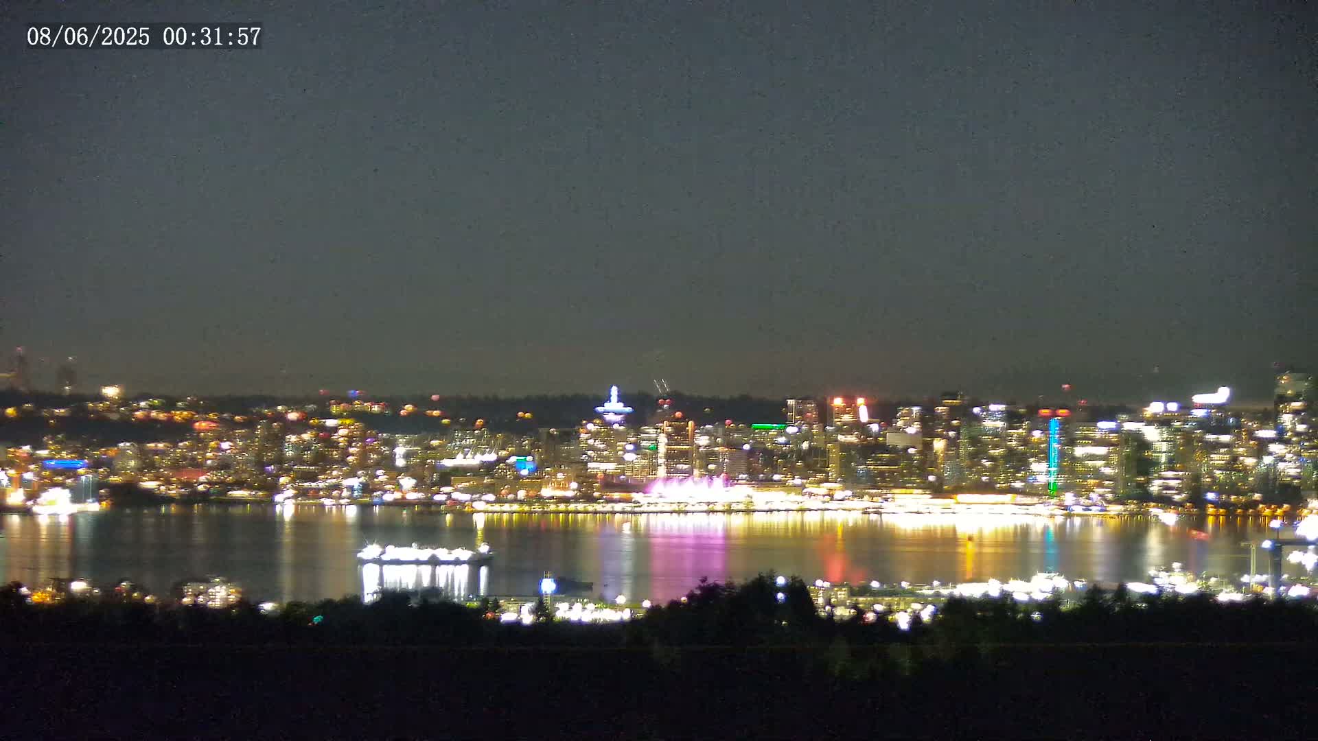 A brightly lit city skyline is reflected in a dark body of water under a night sky.