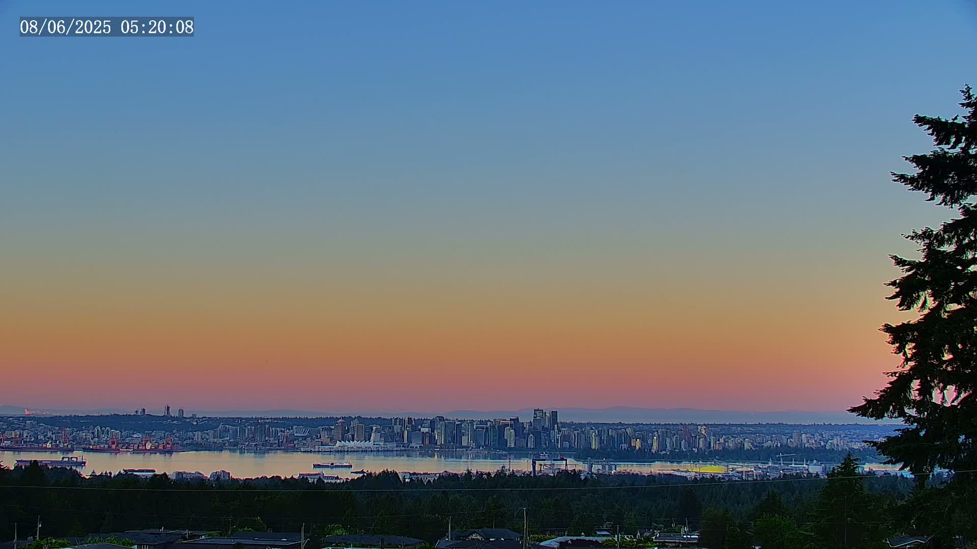 A city skyline is visible across a body of water at sunset, with a clear sky exhibiting a gradient of soft pink and orange hues.