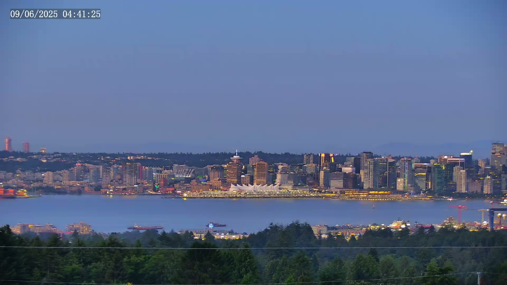 A city skyline at twilight, viewed from across a calm body of water and partially obscured by a foreground of trees under a clear, dusky sky.
