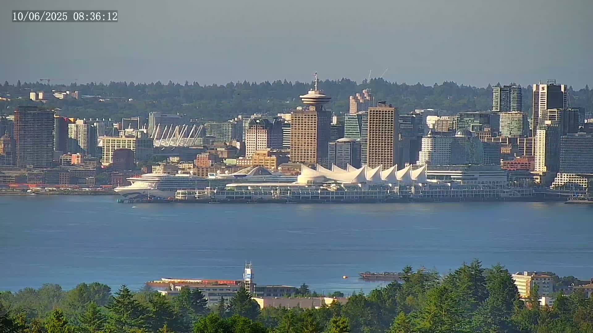 A distant view of a city skyline, featuring a large cruise ship docked near a white-tented building, is seen across a calm body of water, under hazy, sunny conditions.
