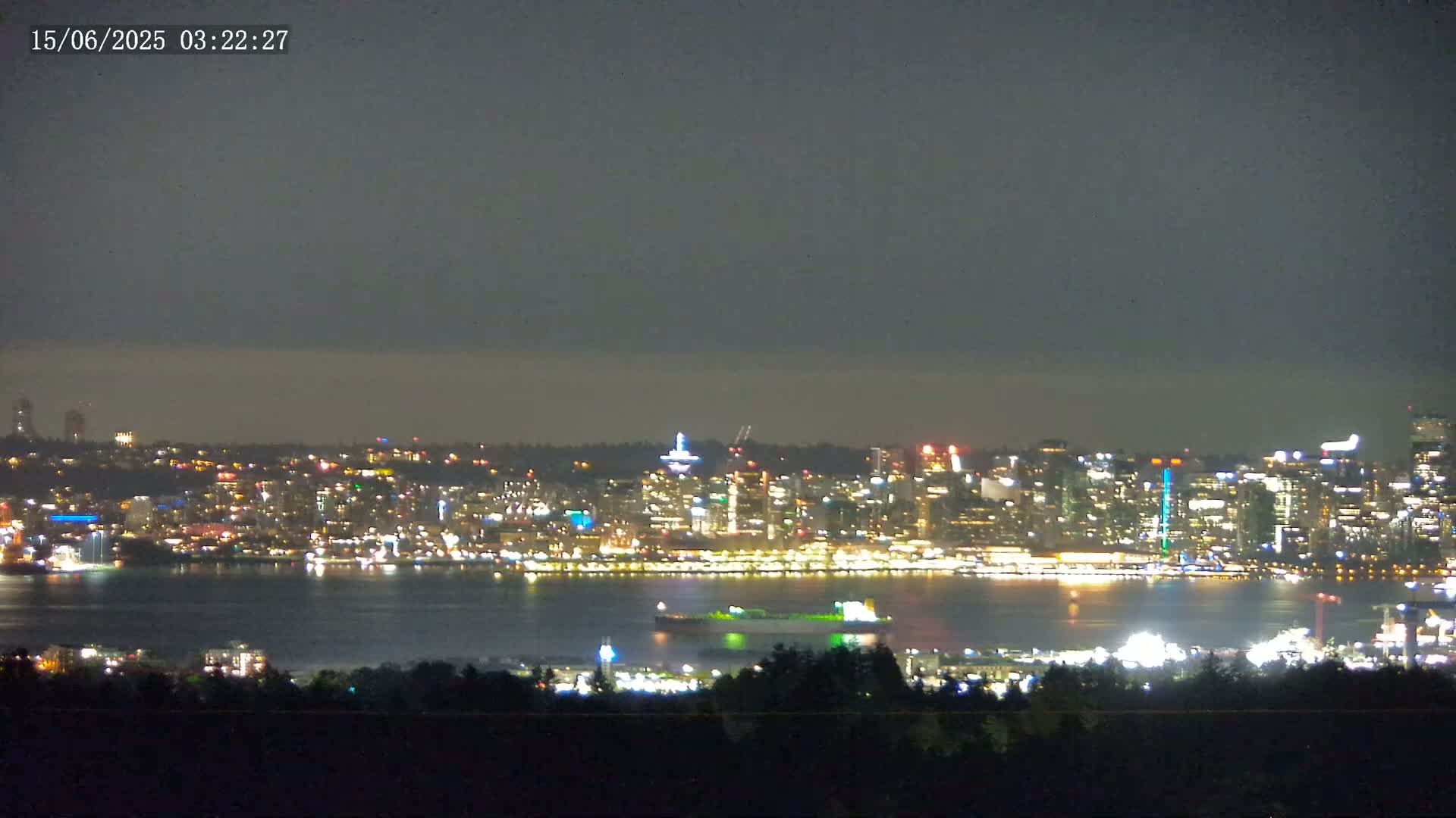 A nighttime view of a brightly lit city skyline across a body of water, under a dark, cloudy sky.