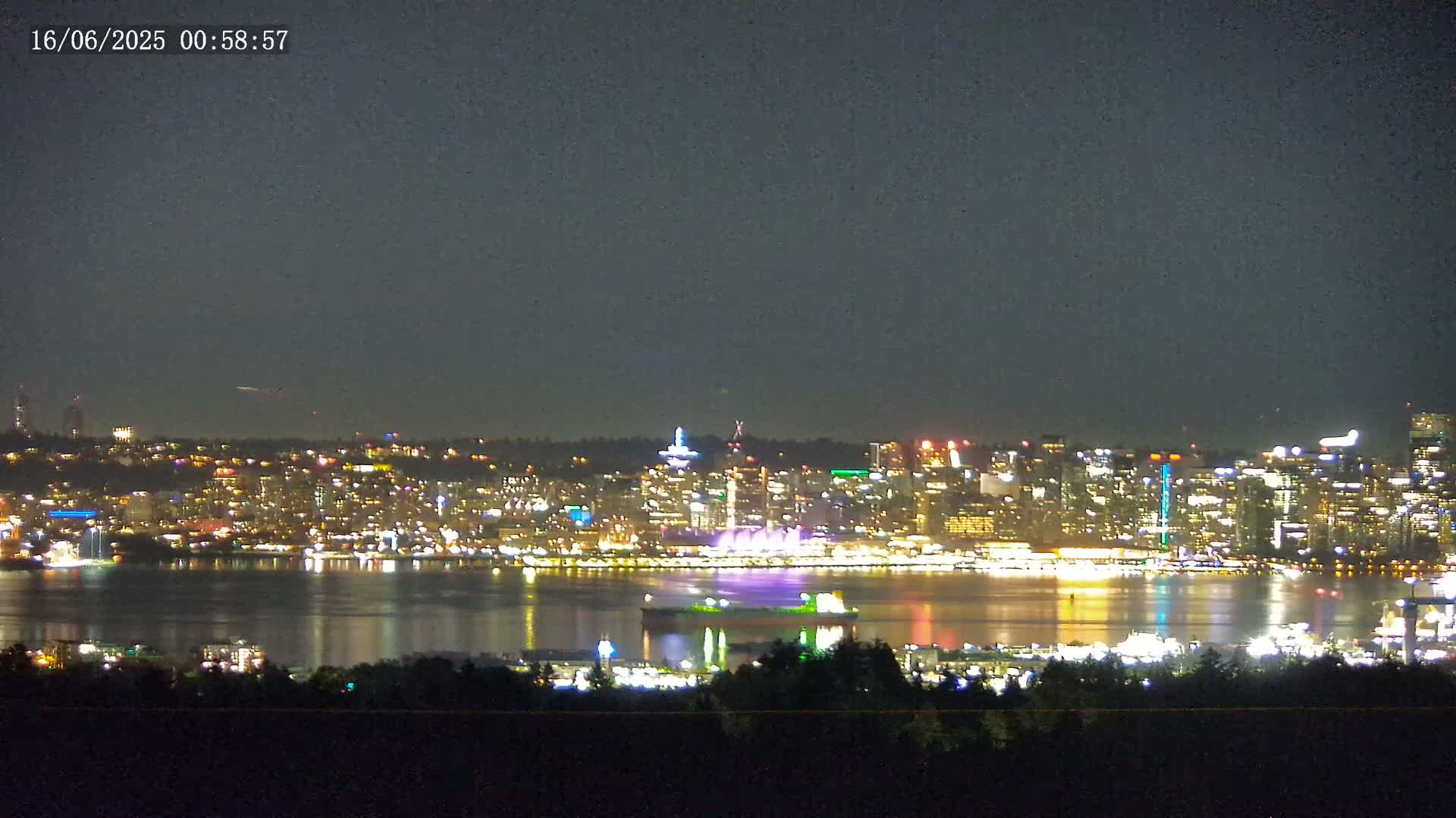 A nighttime view of a brightly lit city skyline across a calm body of water, under a dark, clear sky.