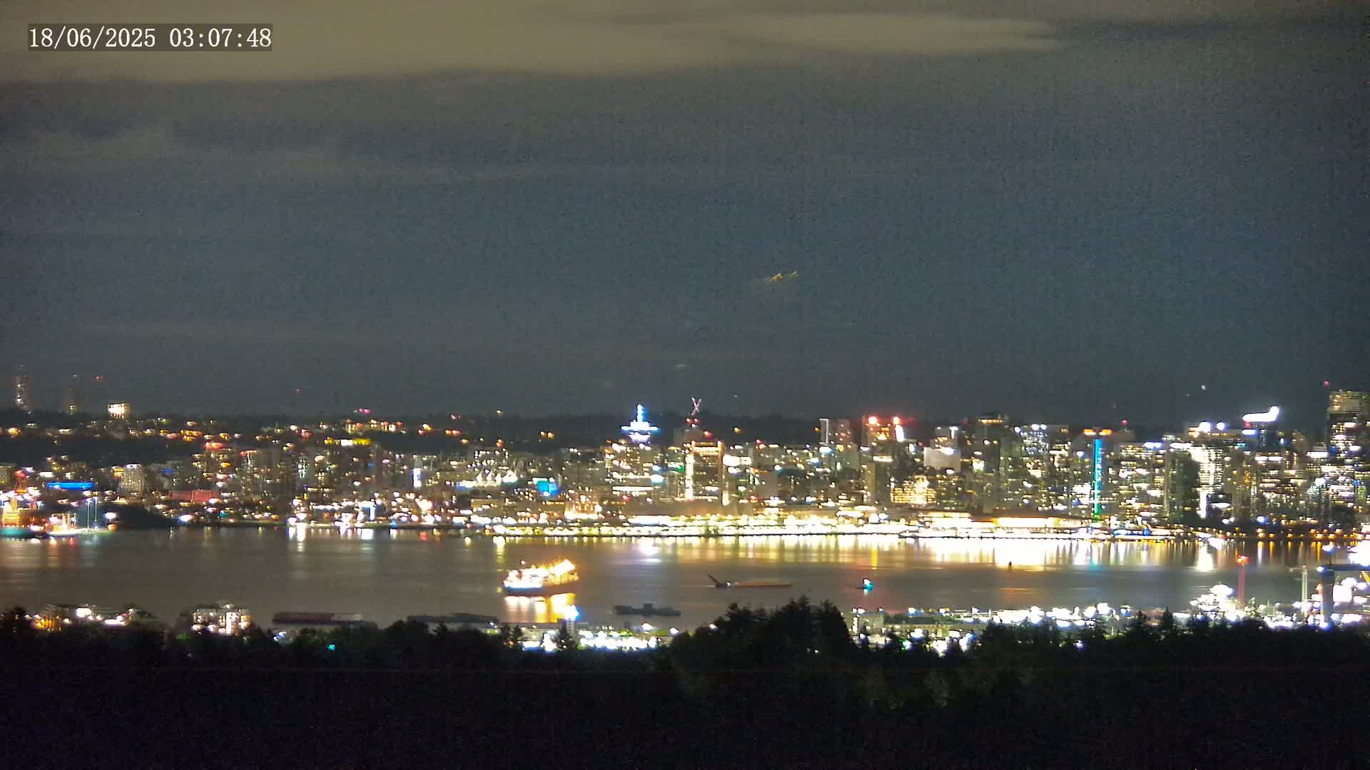 A brightly lit city skyline across a body of water is viewed from a distance at night under a mostly cloudy sky.
