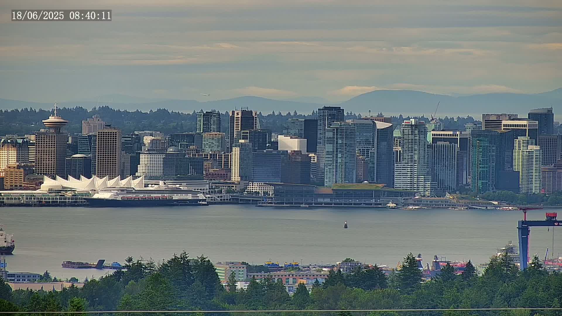 A distant view of a city skyline, with numerous skyscrapers and a large cruise ship docked at the waterfront, on a calm, overcast day, backed by mountains.