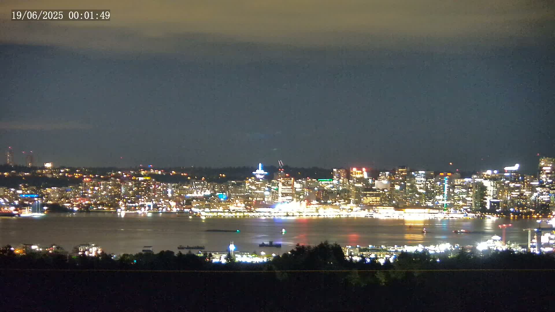 A brightly lit city skyline across a dark body of water is visible at night under a cloudy sky.