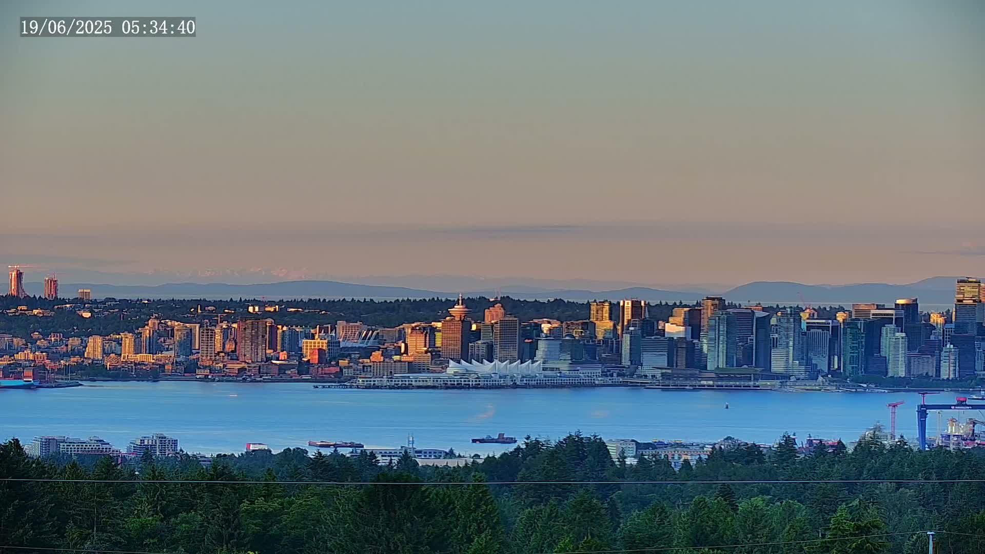 A city skyline, including skyscrapers and a large convention center, sits on the water's edge beneath a pale pink and orange sunset sky, with mountains visible in the distance.