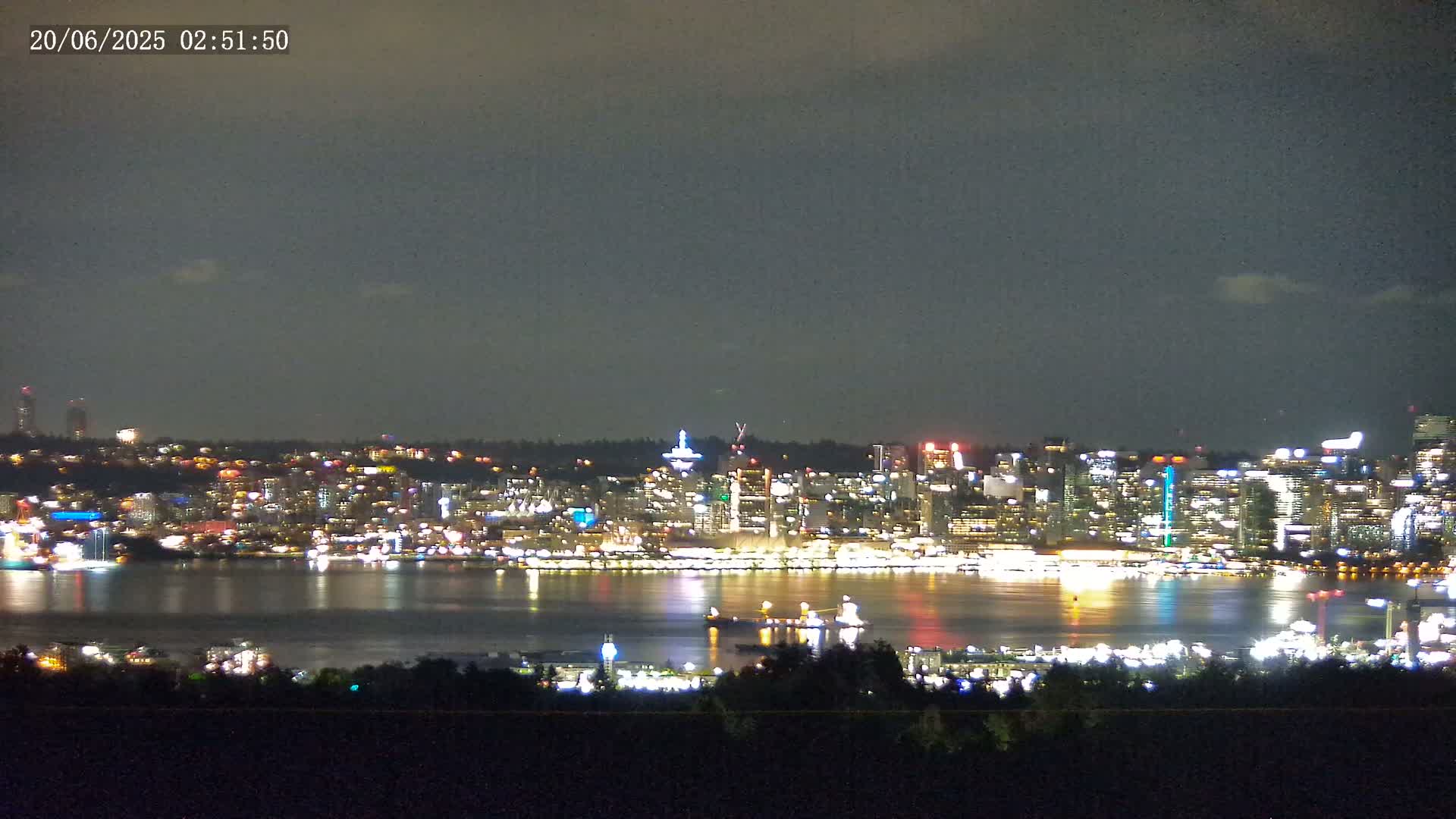 A brightly lit city skyline at night is seen across a dark body of water under a mostly cloudy sky.