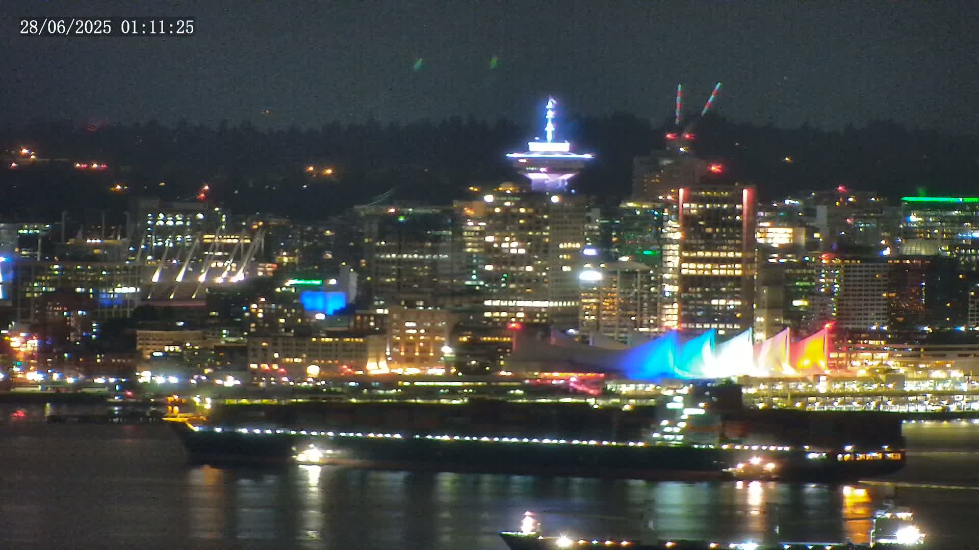 A brightly lit city skyline at night is seen across a dark body of water, with a large ship in the foreground.