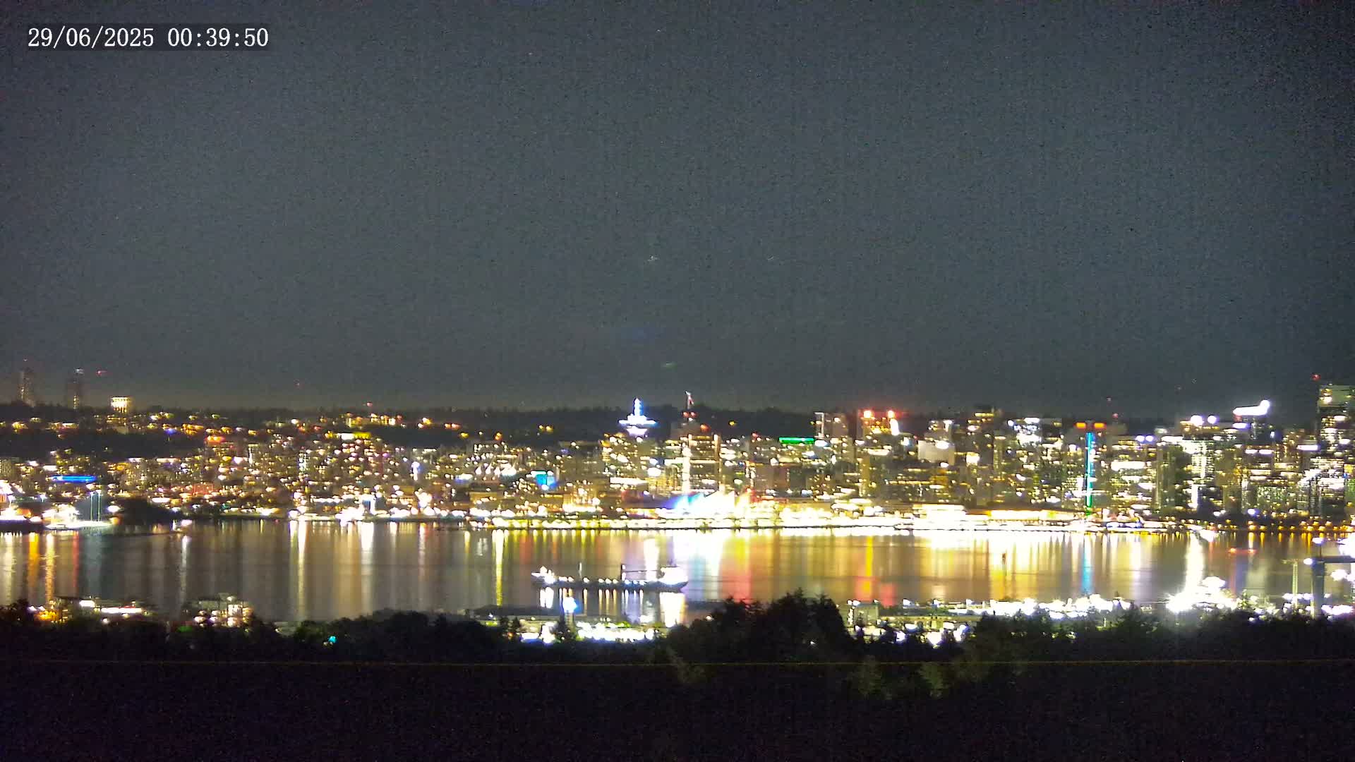 A brightly lit city skyline is reflected on a calm body of water under a dark night sky.