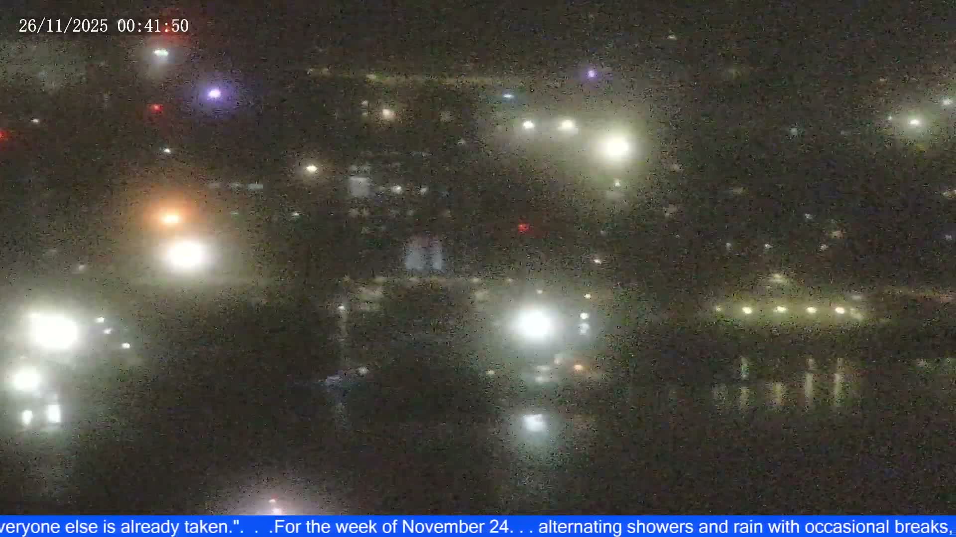 The image displays a very grainy and blurry outdoor night scene illuminated by numerous diffused lights, some reflecting on what appears to be water, under rainy or showery conditions.