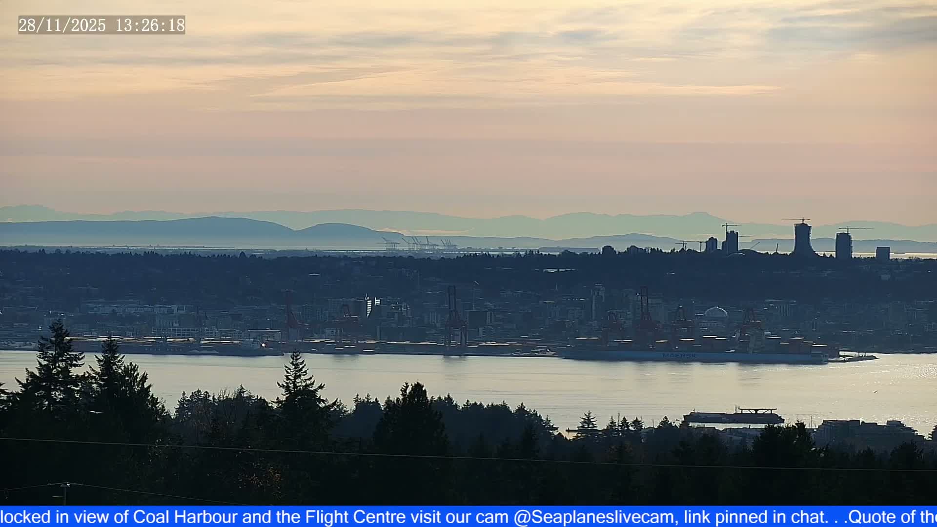 An expansive outdoor view reveals a calm harbor with a cargo ship and an industrial city skyline, backed by hazy mountains under an overcast sky, with dark evergreen trees framing the foreground.