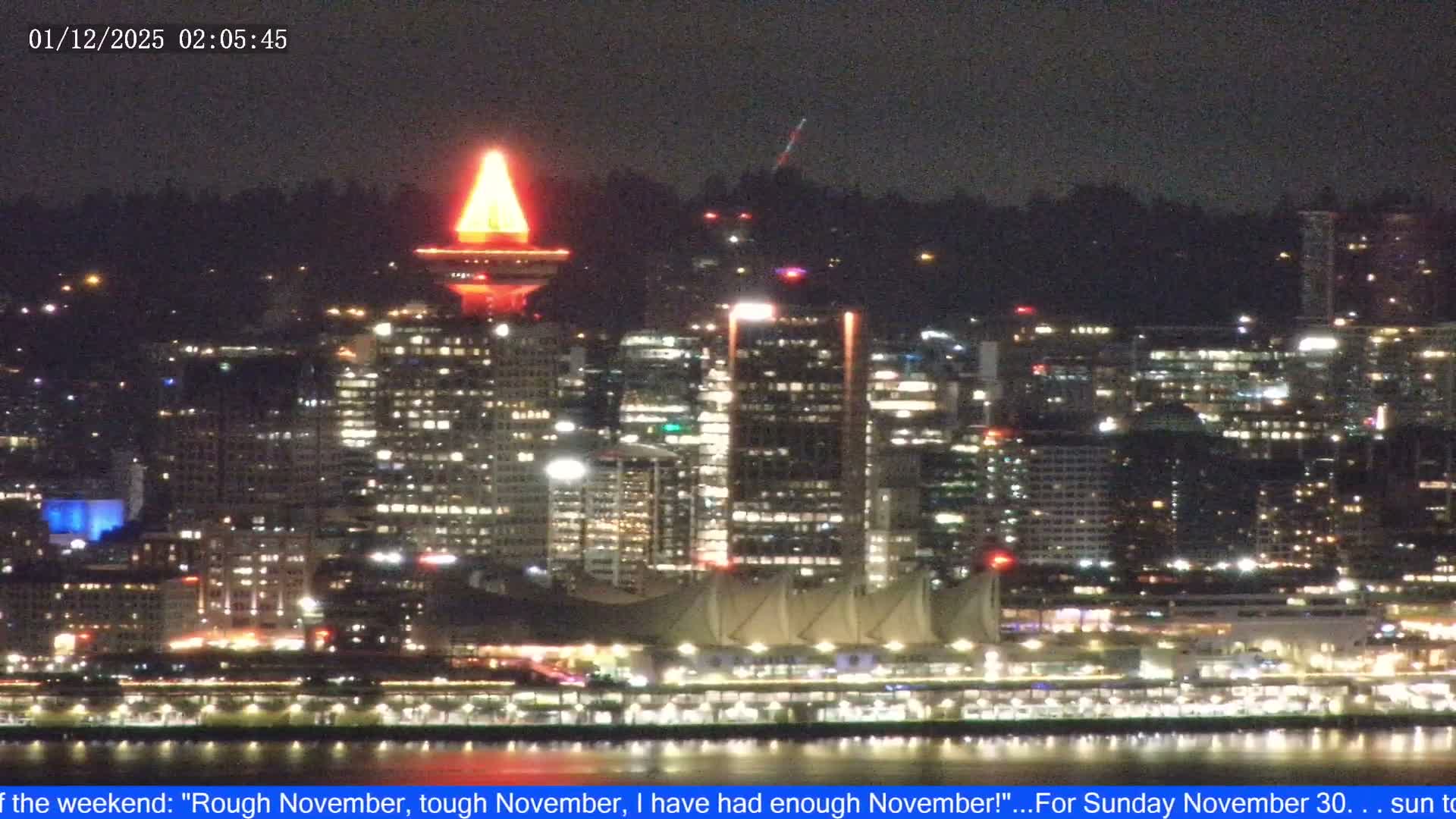 A vibrant city skyline, featuring a prominently illuminated red and orange conical tower and buildings along a waterfront reflecting lights on the water, is seen against a dark, clear night sky.