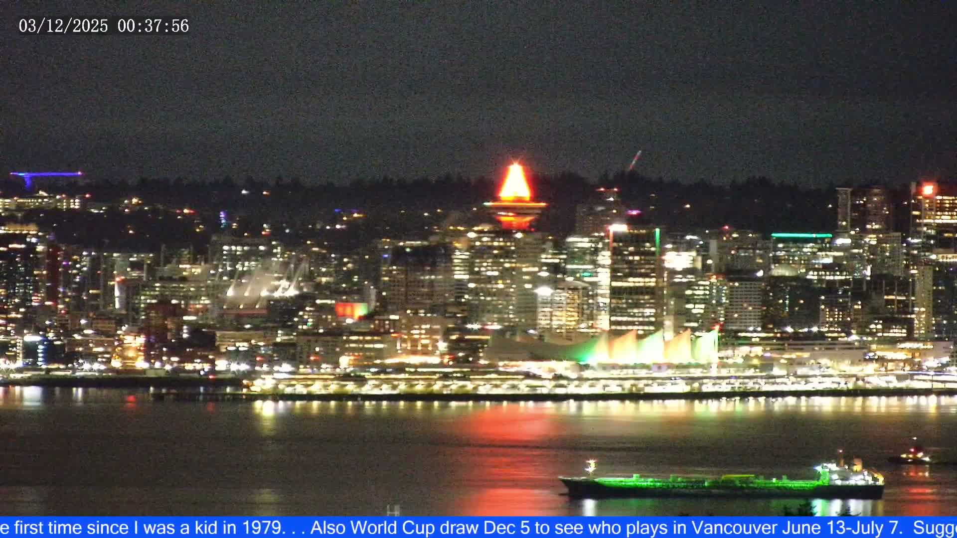 Vibrant city lights illuminate a clear night skyline, featuring a prominently glowing red-topped tower, various buildings, and colorful reflections on the dark water where a green-lit ship sails.