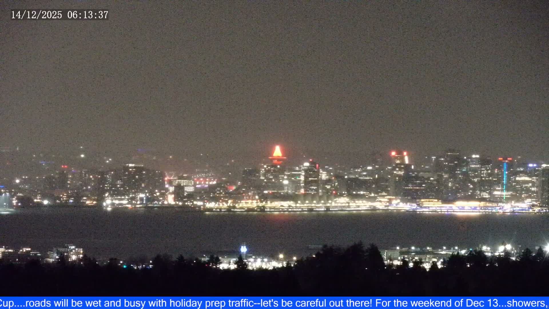 Vibrant city lights illuminate a clear night skyline, featuring a prominently glowing red-topped tower, various buildings, and colorful reflections on the dark water where a green-lit ship sails.
