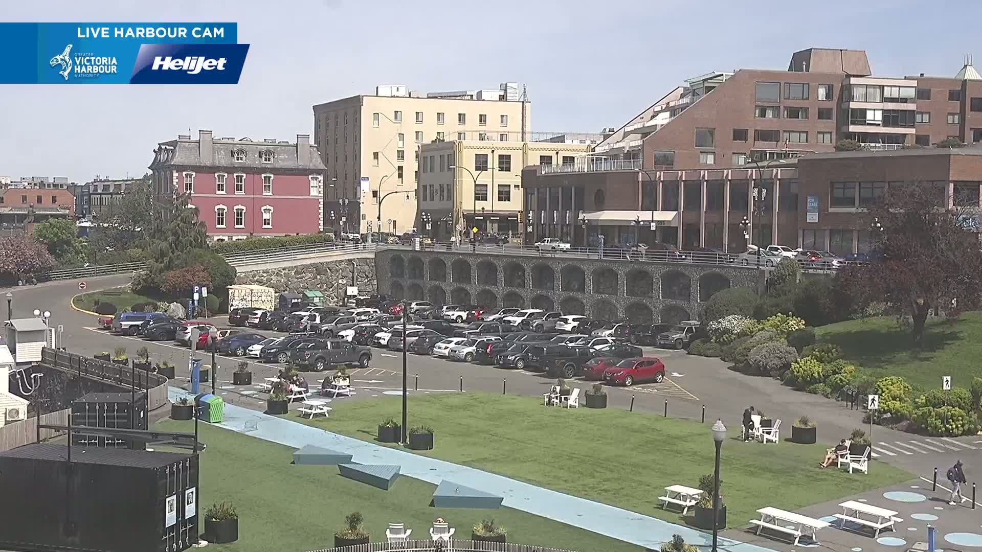 A sunny day shows a waterfront parking lot full of cars, adjacent to a grassy area with people sitting at picnic tables, all situated in front of several multi-story buildings.