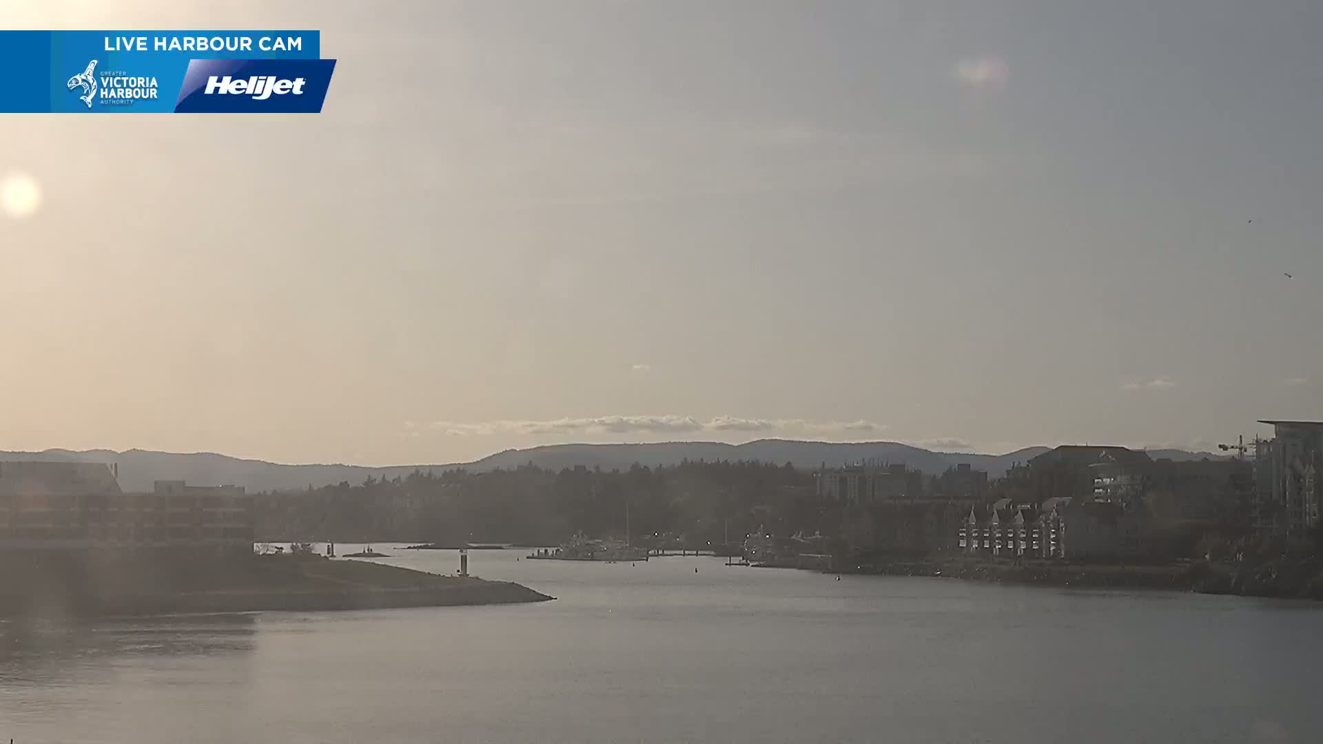 A hazy, sunny day reveals a harbor scene with calm water, boats docked, buildings along the shore, and distant mountains.
