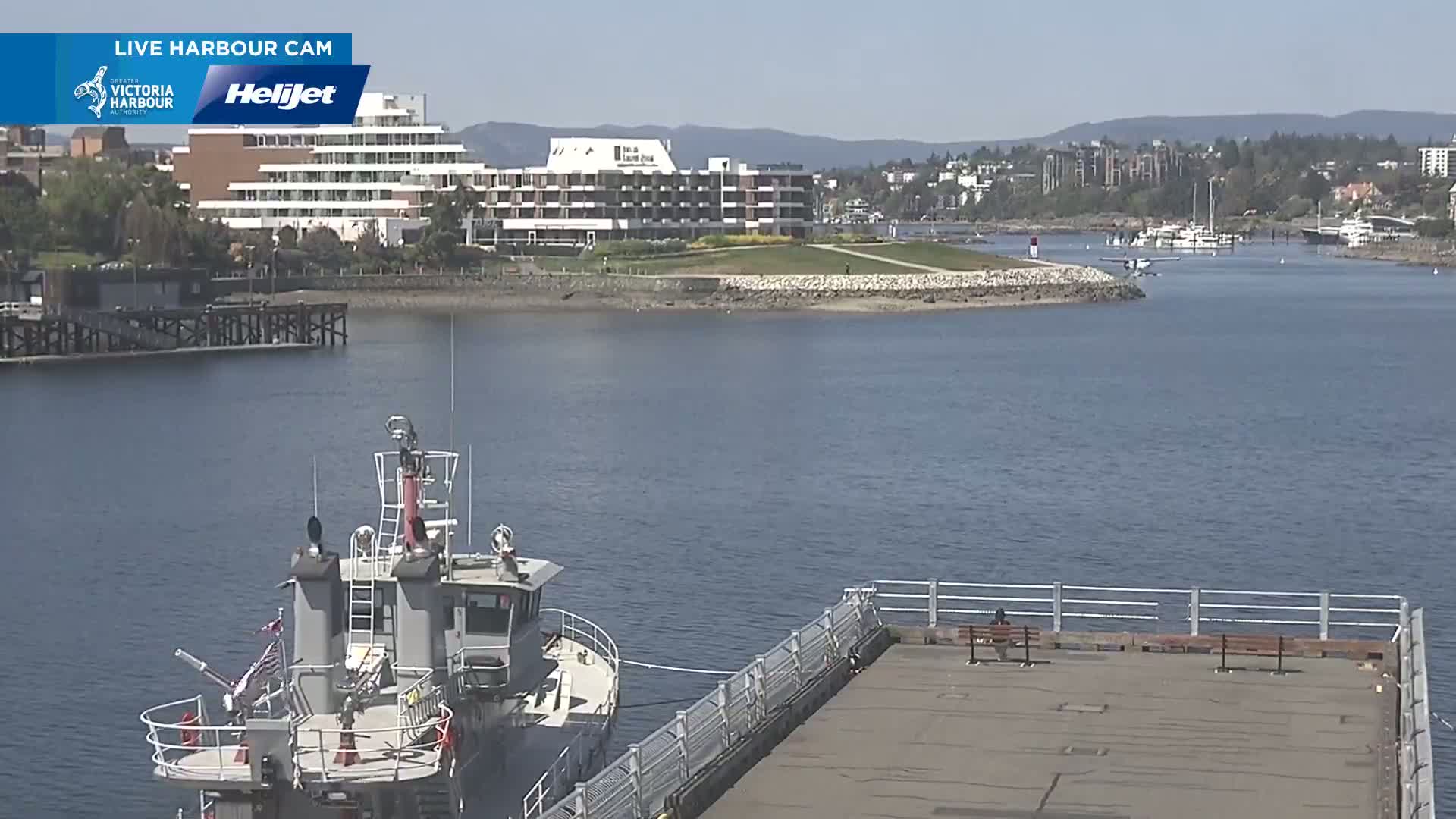 A gray harbor boat is docked at a pier next to calm blue water, with a large hotel and residential buildings visible across the water under a sunny sky.