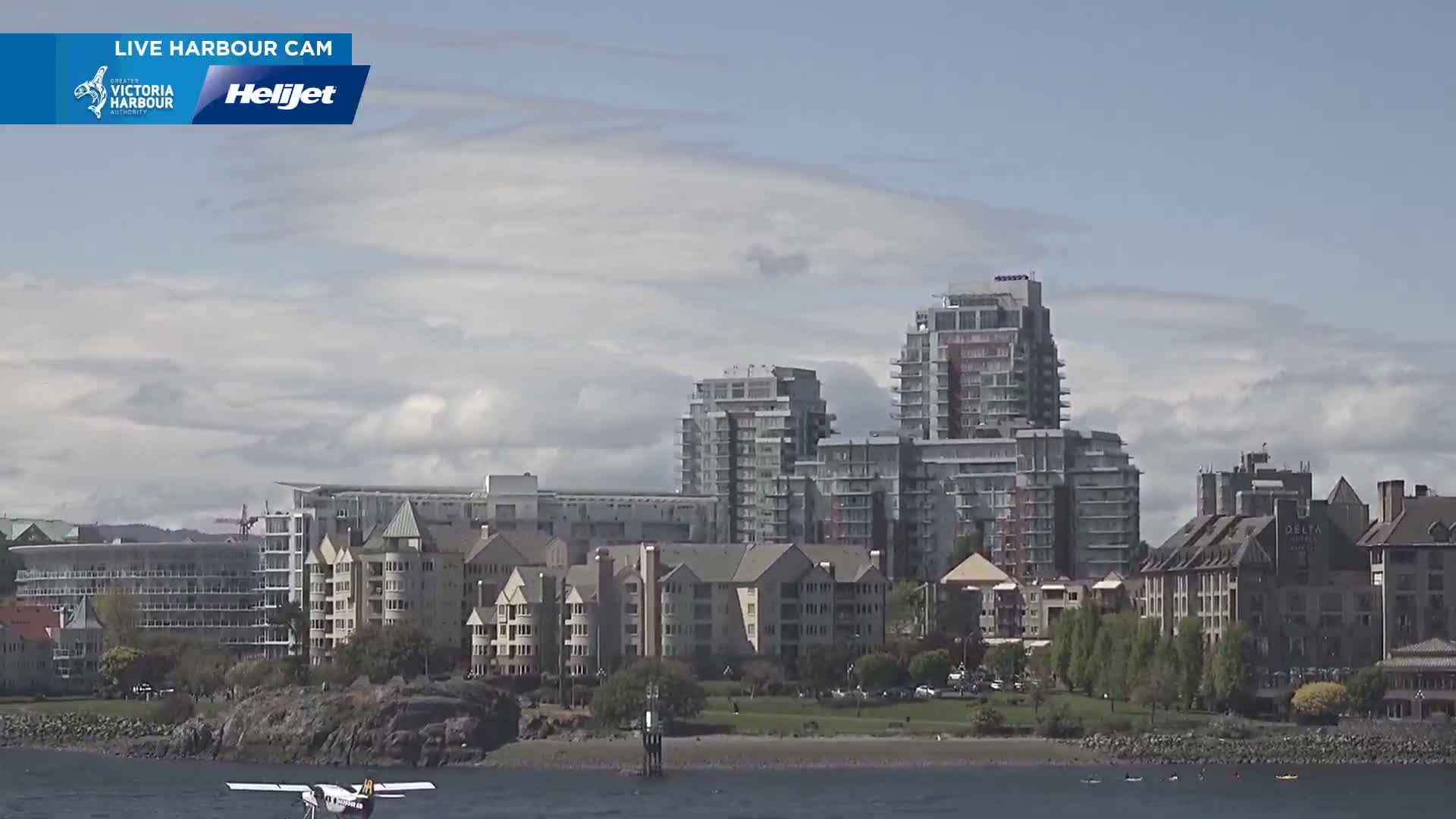 A seaplane is on the water in front of a waterfront cityscape under a partly cloudy sky.