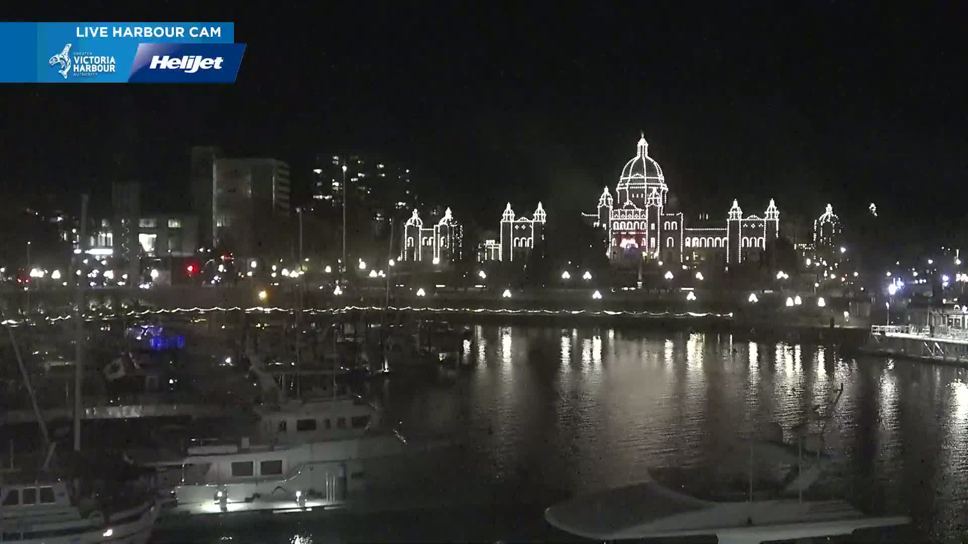 At night, a large building is illuminated with white lights across its exterior, reflecting in the calm water of a harbor filled with boats.