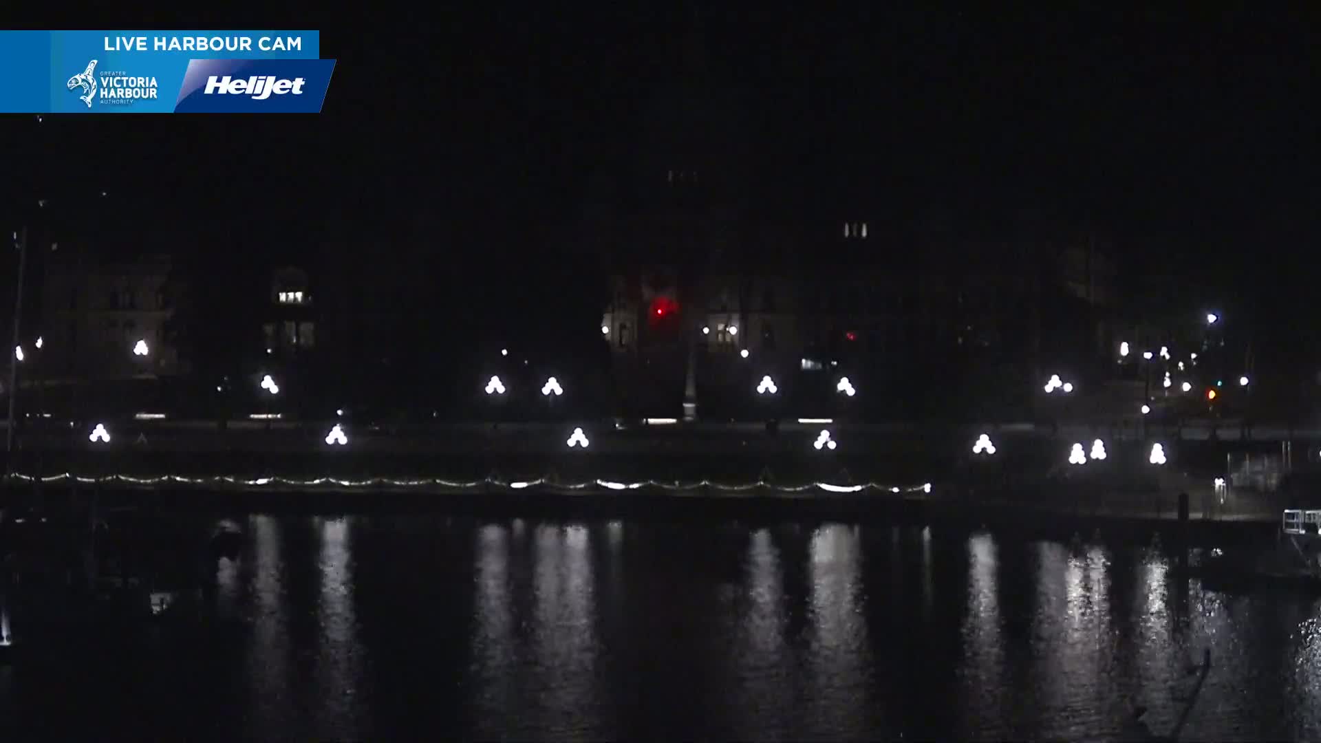 A nighttime view of a calm harbor with lights reflecting on the water, illuminating a waterfront area with buildings in the background.