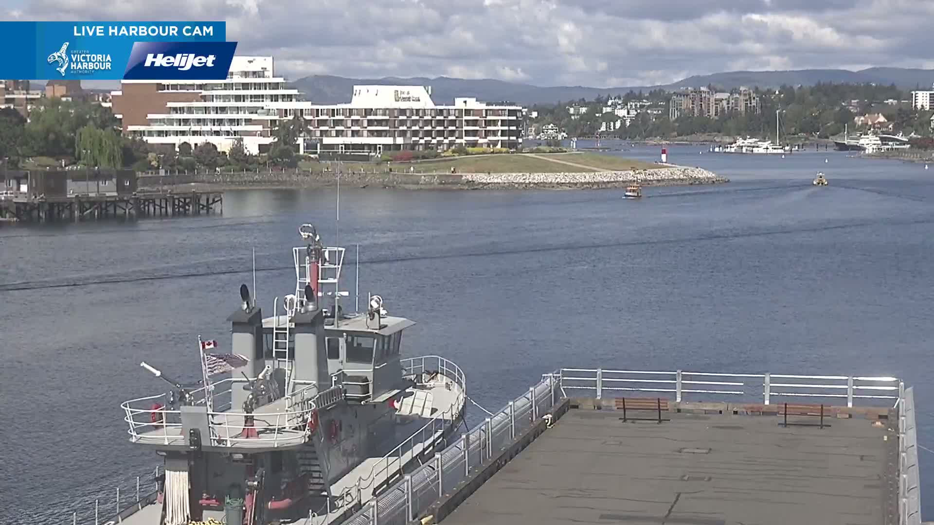 A harbor scene on a partly cloudy day shows a tugboat docked next to a pier, with a large hotel and other buildings visible in the distance across the water.