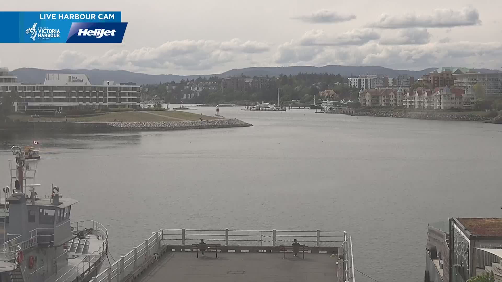 A calm, overcast day shows a harbor with a few boats, a waterfront building, and residential buildings across the water.
