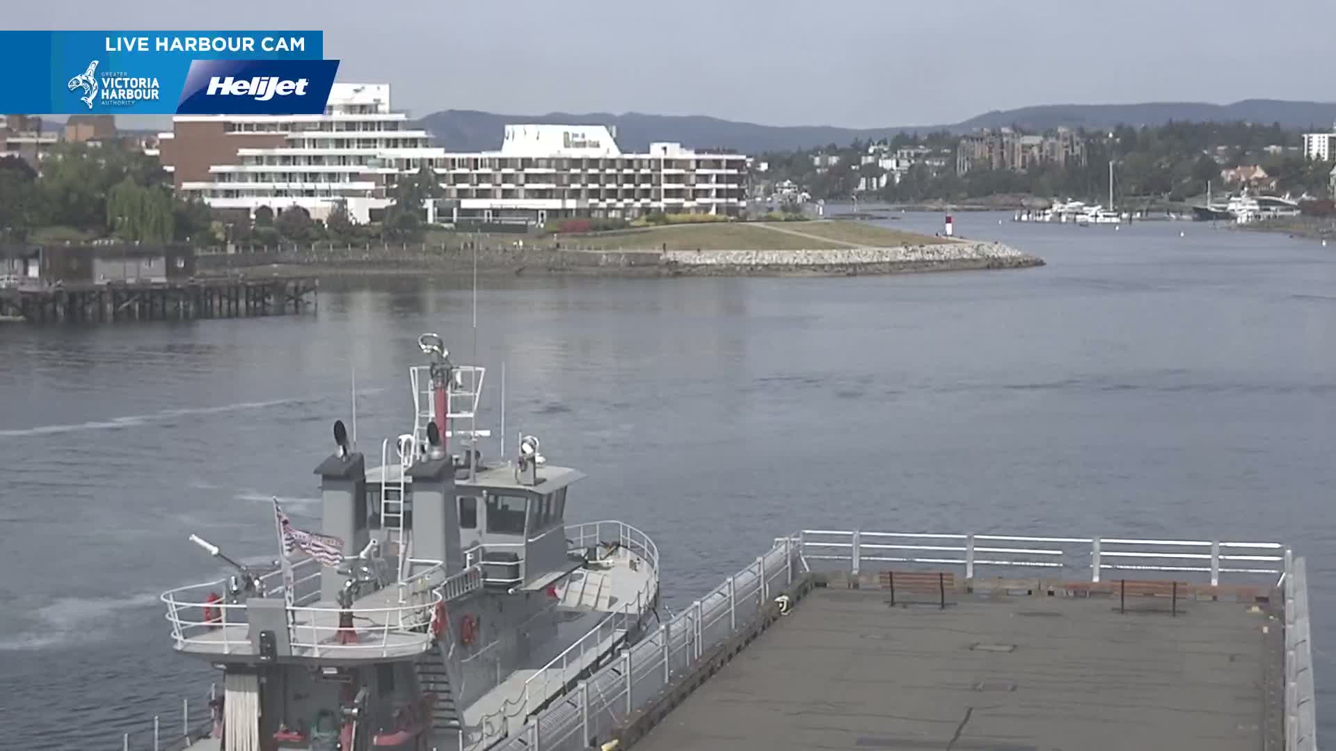 A grey fireboat is moored near a dock next to calm water, with a hotel and residential buildings visible in the background on a partly cloudy day.