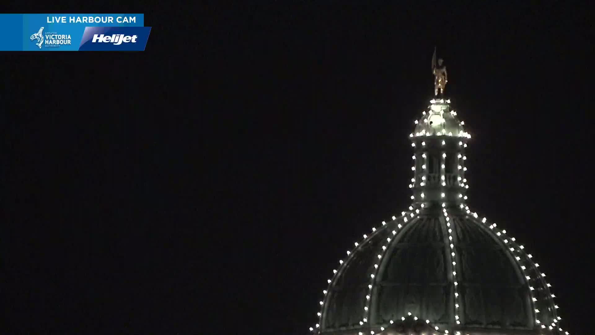 A dark nighttime image shows the top of a dome-shaped building, adorned with small white lights and topped with a statue.