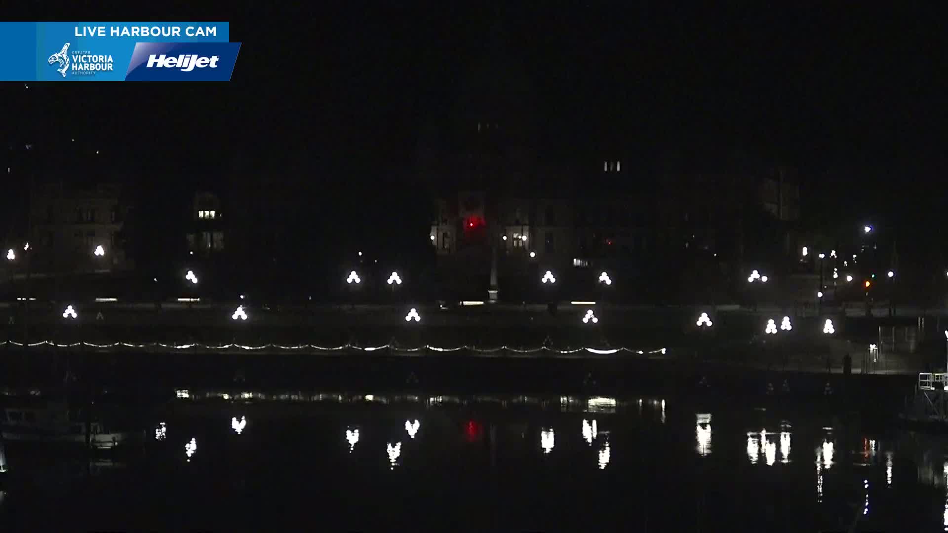 A dark nighttime scene shows a body of water reflecting the lights of a waterfront building and promenade.
