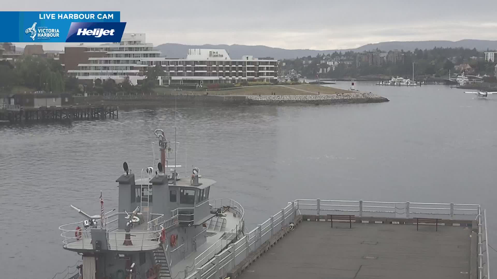 A gray tugboat is moored near a dock in a harbor on an overcast day, with a large hotel and residential buildings visible in the background.